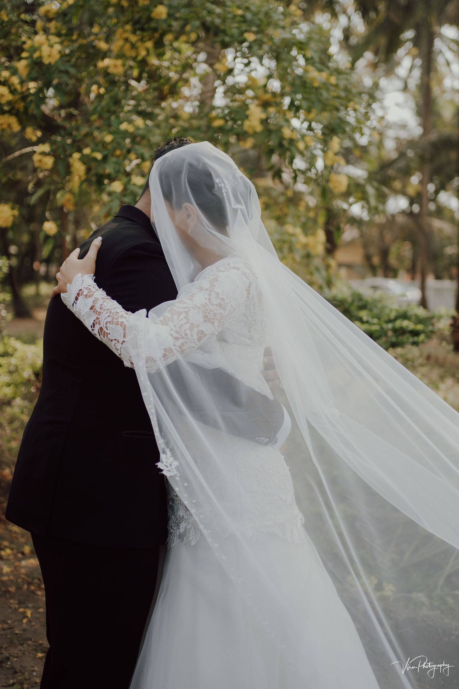 A couple embraces during their wedding ceremony outdoors with a flowing white veil and lace dress against autumn foliage.
