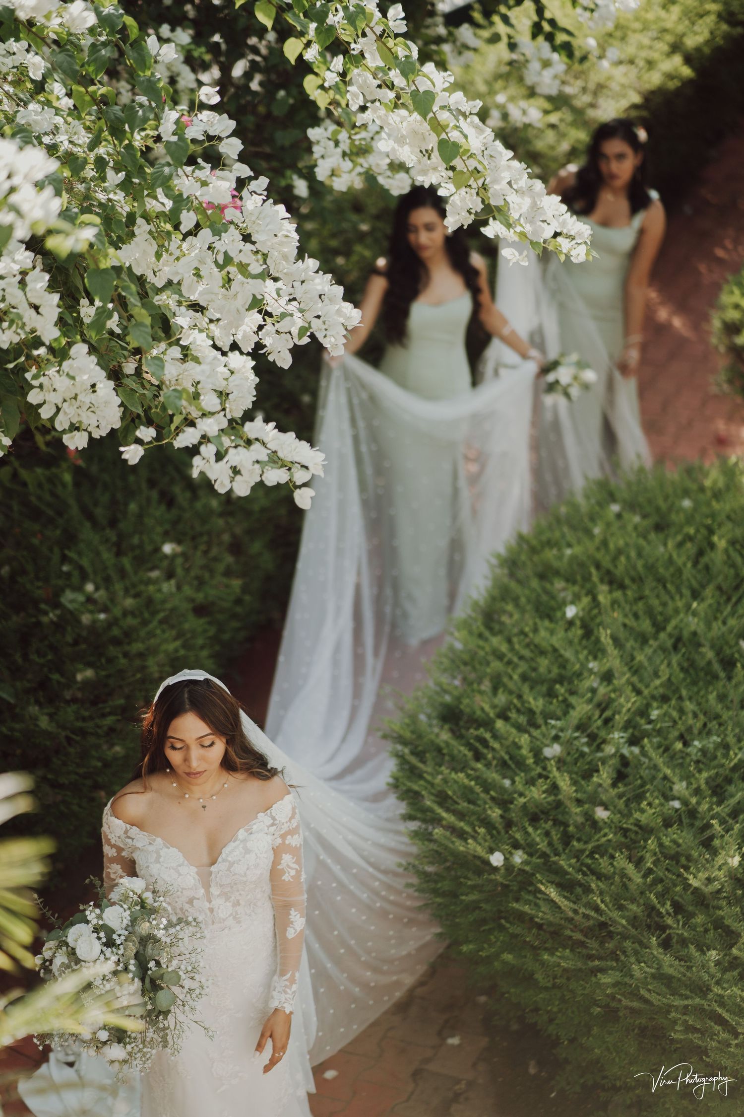 A romantic wedding scene with ethereal white gowns and flowing veils amid blooming florals and lush greenery.