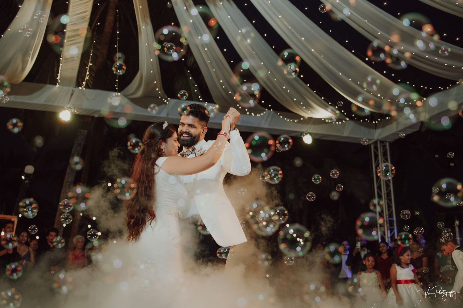 Newlyweds share their first dance surrounded by floating bubbles and fog on a romantic dance floor under draped lights.