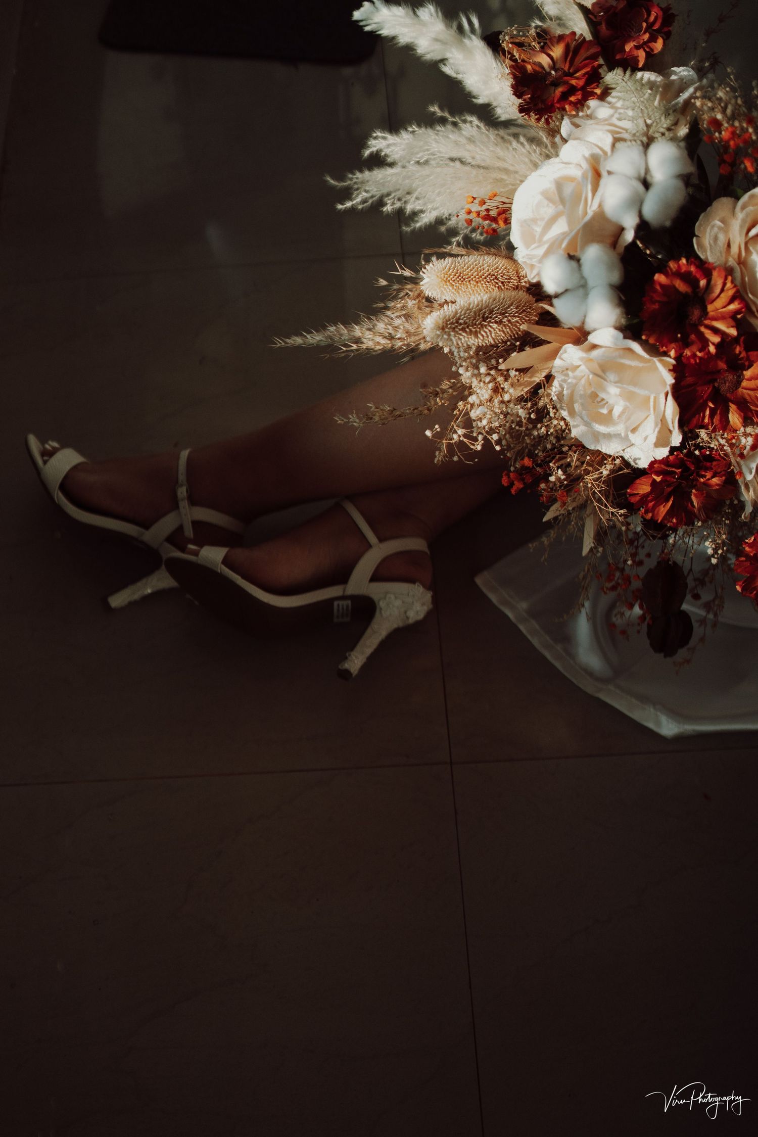 Moody floral arrangement with pale roses, pampas grass and burgundy blooms against dark background in dramatic lighting.