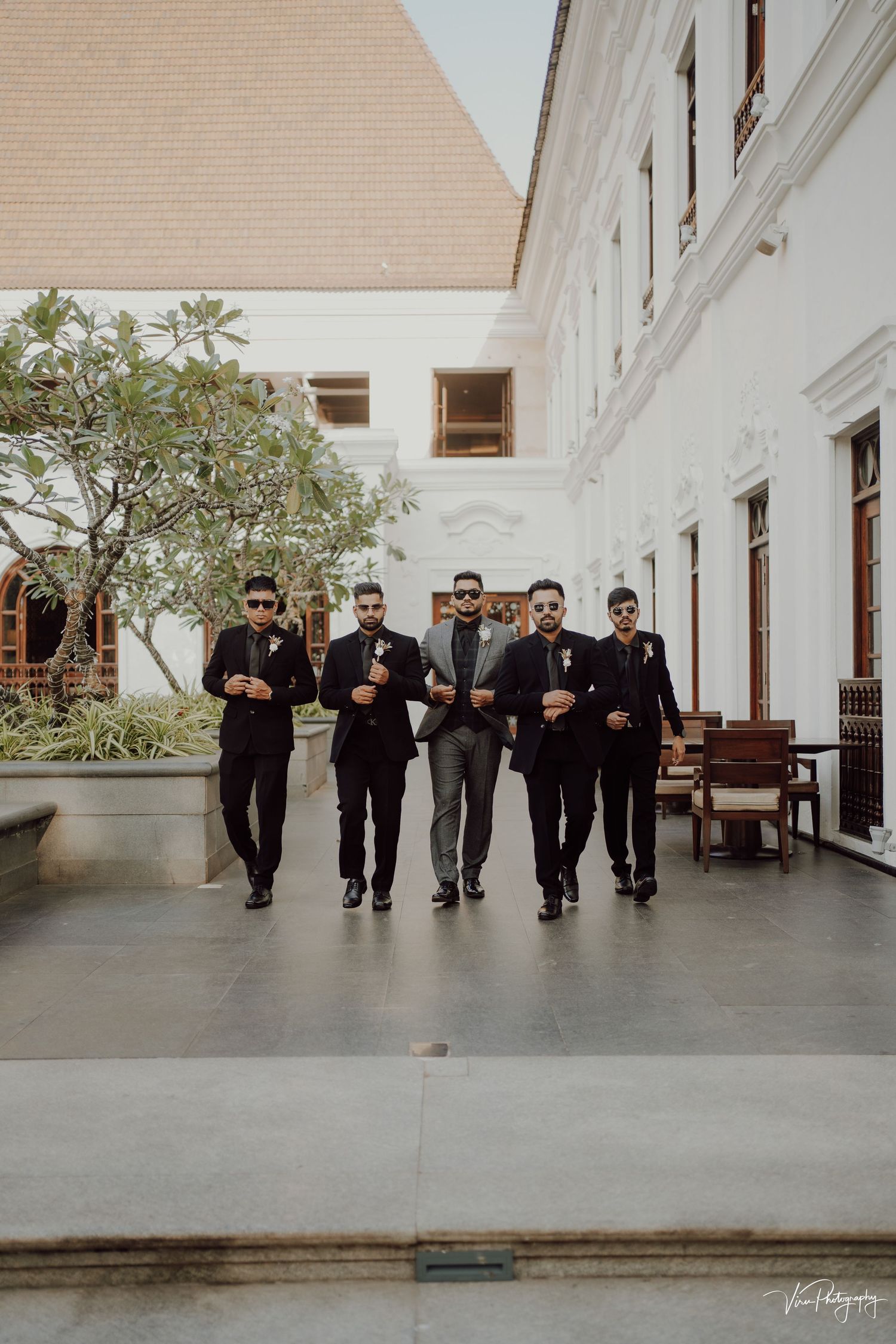 A group of groomsmen in black suits walk together down an elegant covered walkway at a wedding venue.
