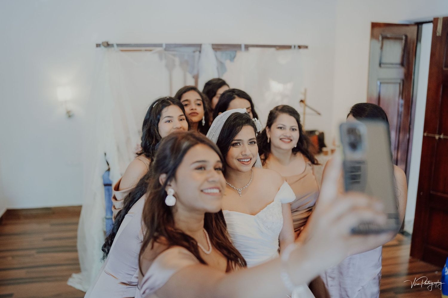 A group in white dresses take a selfie together while smiling and laughing indoors.
