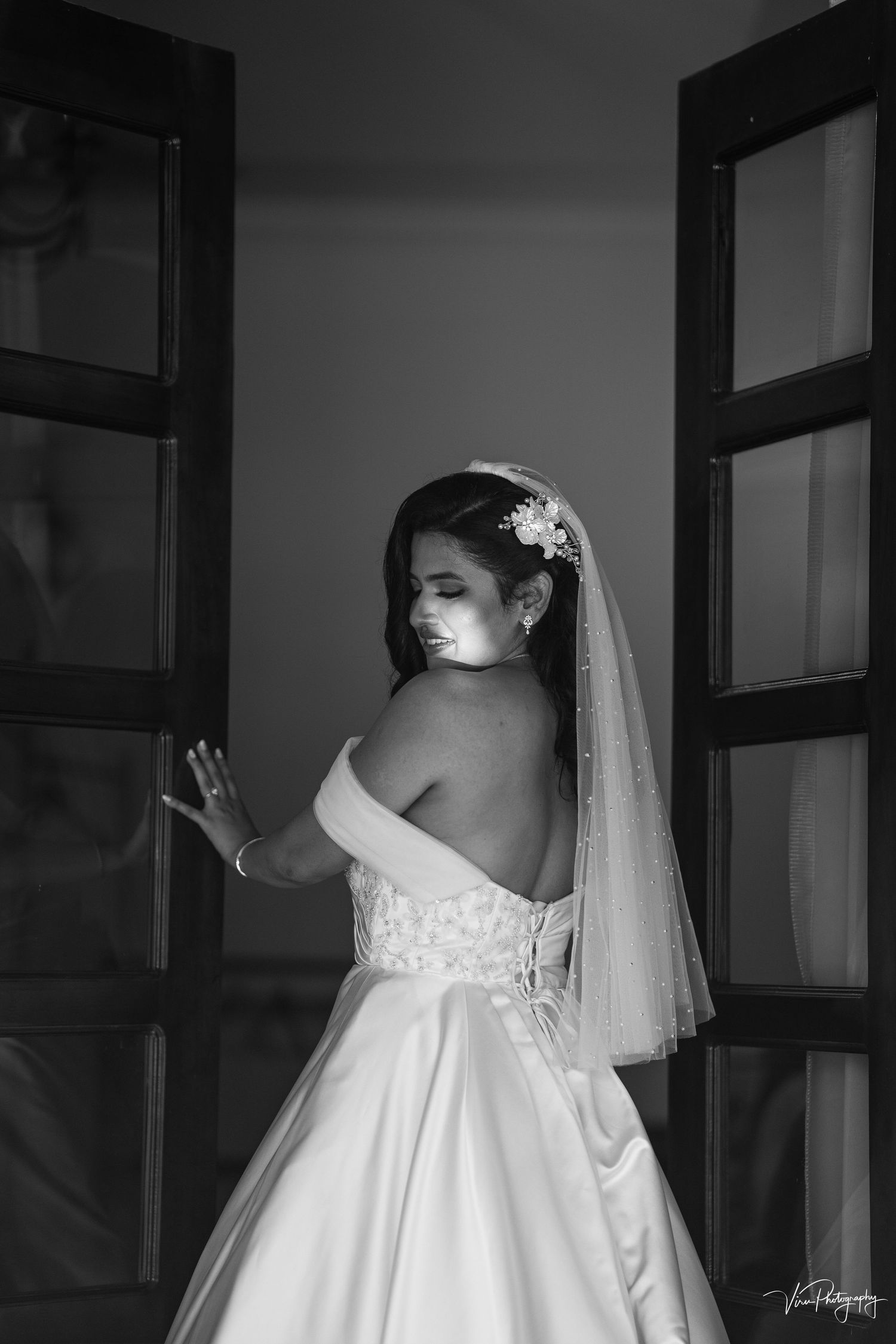A black and white bridal portrait featuring an elegant strapless wedding gown and flowing veil against a windowed backdrop.
