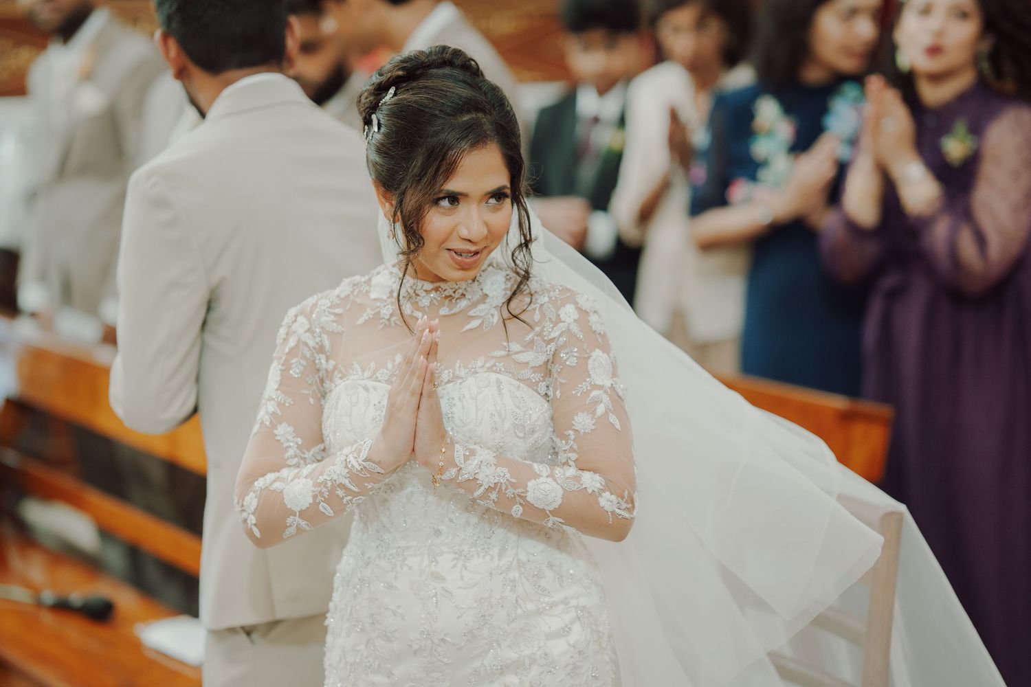 Smiling bride in a lace wedding gown and long veil holds her hands together in prayer during a church ceremony with guests behind her.