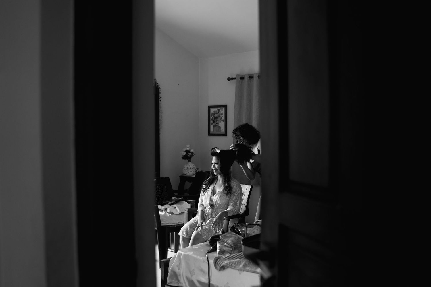 Black and white photo of a bride in a white robe seated while a stylist prepares her hair, seen through a doorway in a softly lit room.