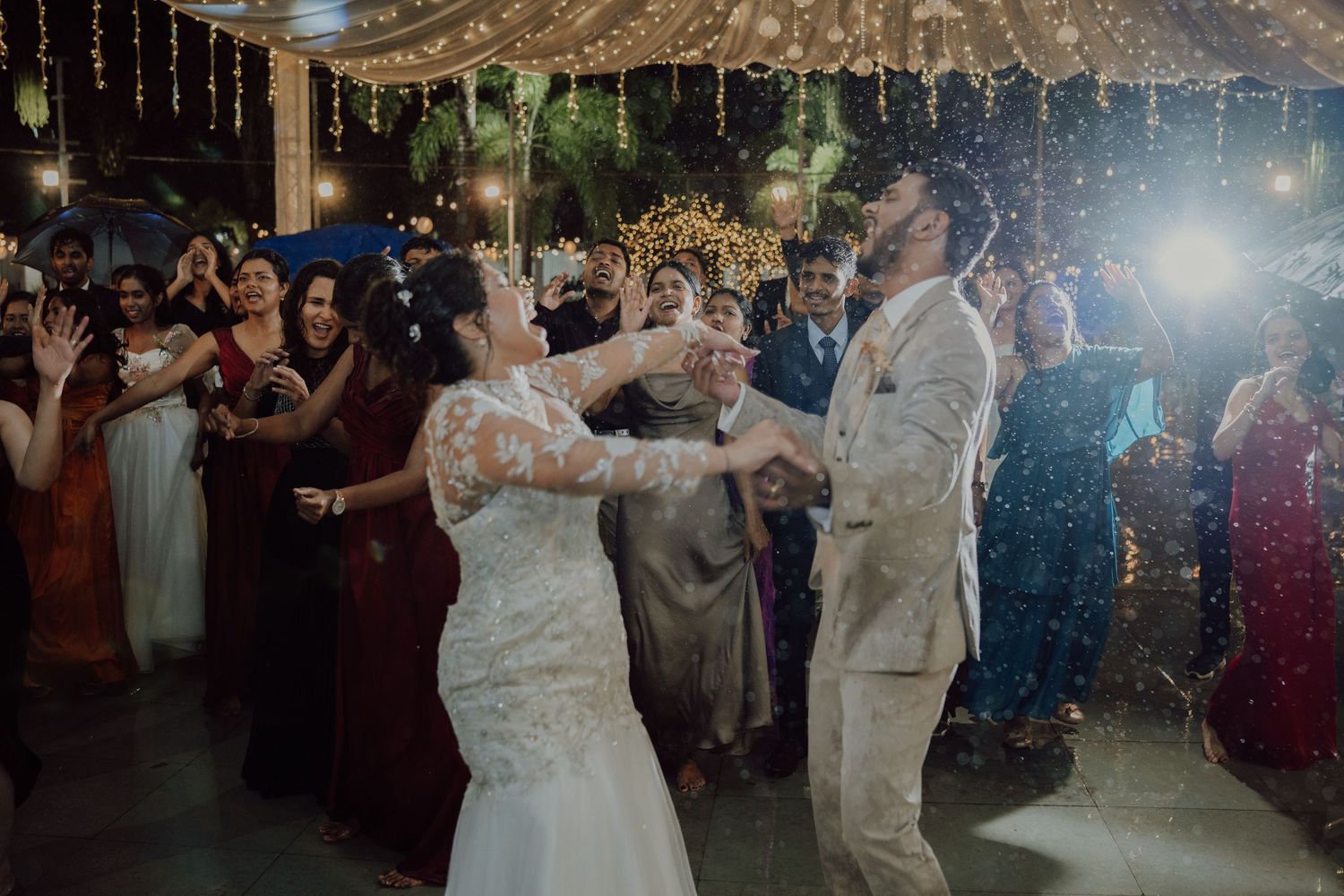 Bride and groom dance with cheering guests under string lights and falling sparkles at an outdoor wedding reception.