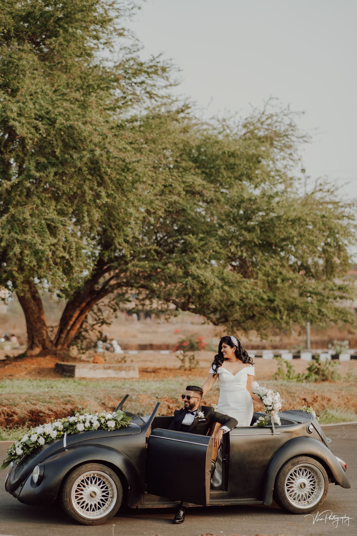 A couple poses with their dog in a vintage Volkswagen beetle convertible under a sprawling tree for wedding photos.