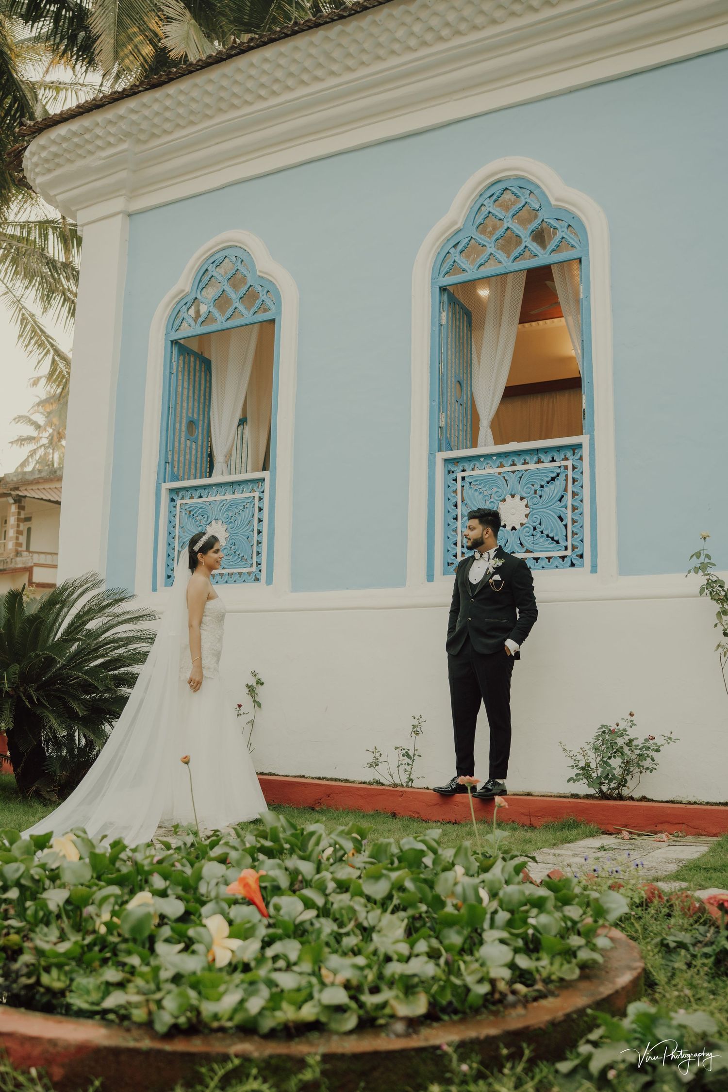Couple poses in front of a vintage blue building with ornate arched windows and tropical foliage in the foreground.