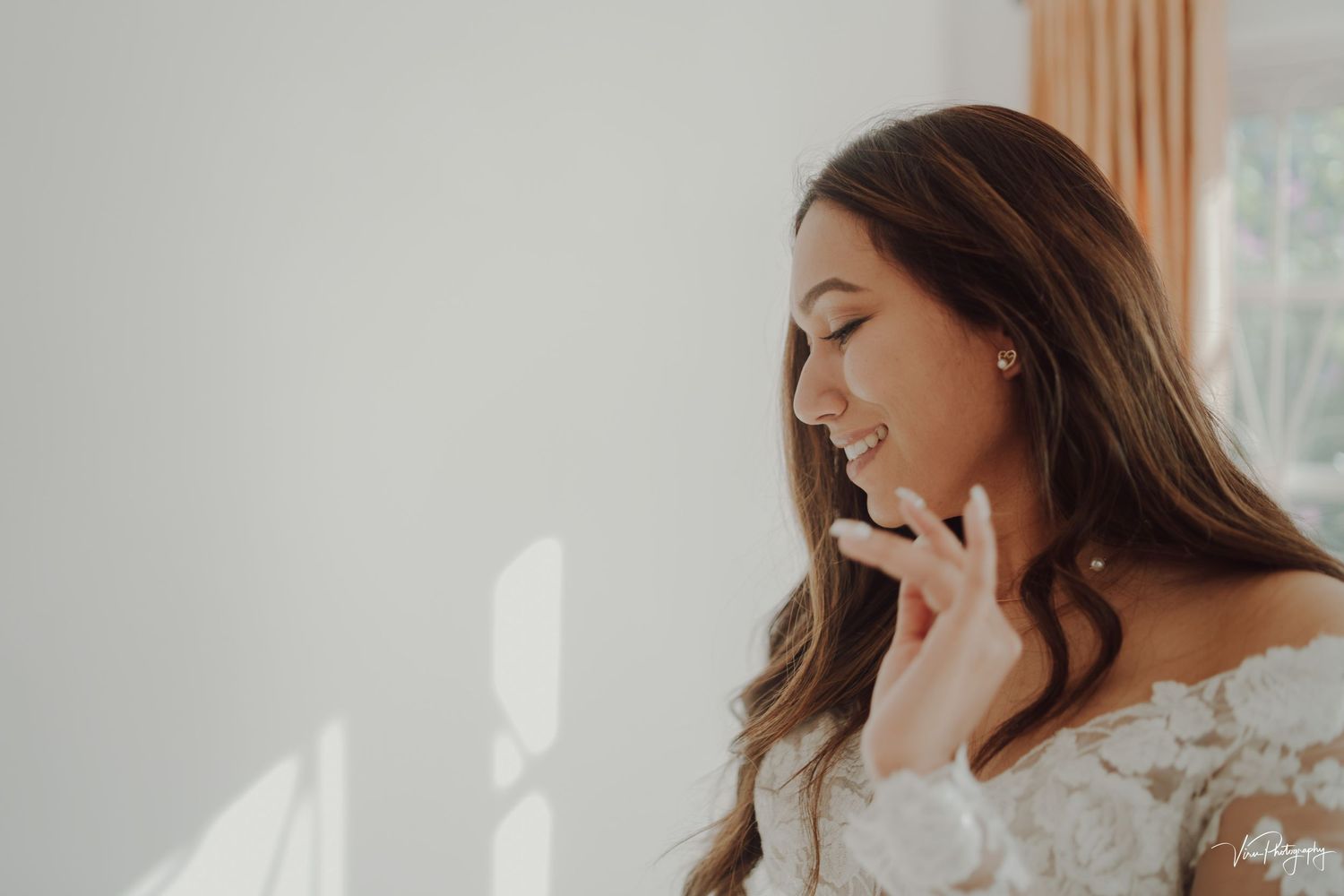 A bride wearing a white lace gown brushes her fingers along her neck while standing in soft window light.