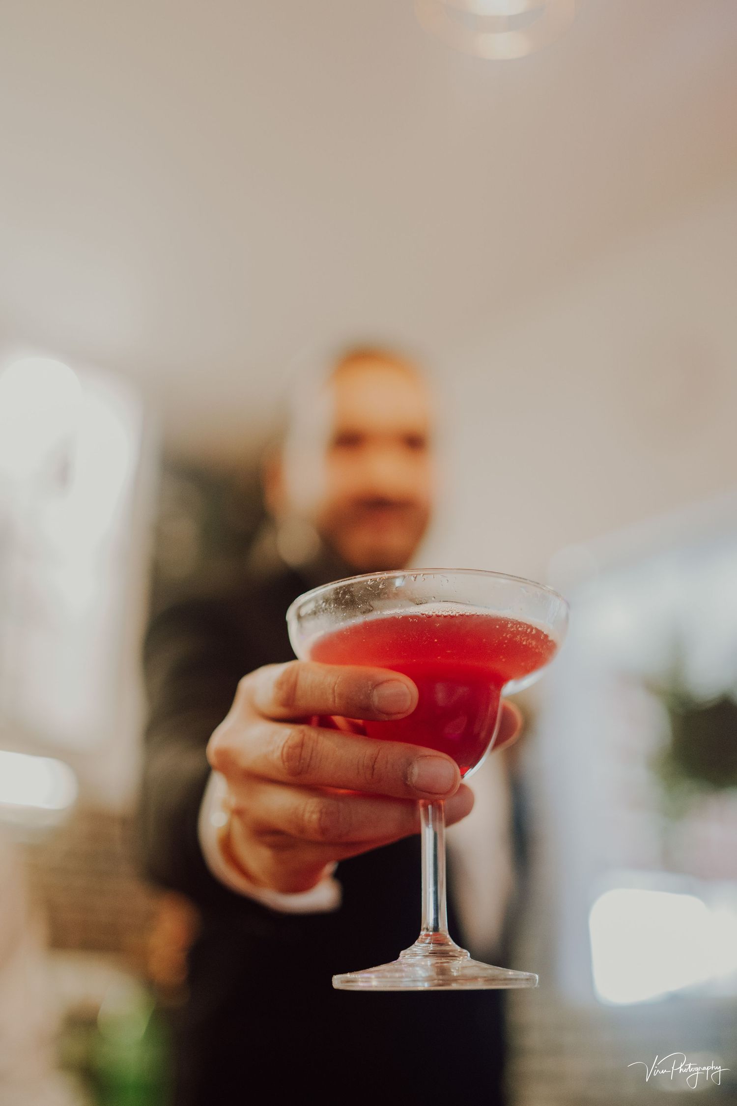 Someone in formal black attire holds a stemmed cocktail glass with vibrant red liquid against a soft-focused background.