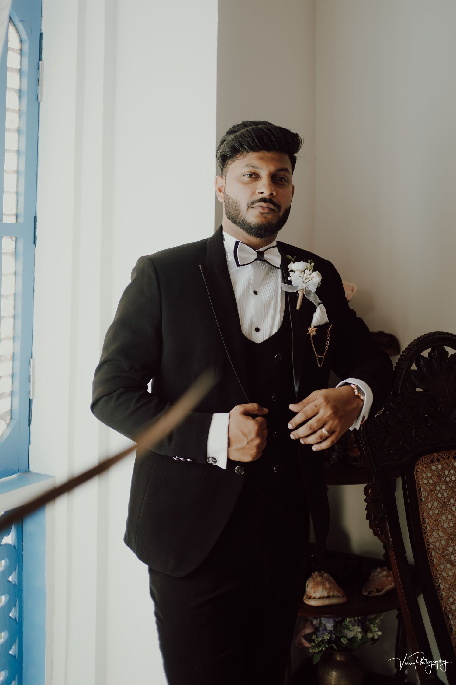 A groom in a formal black tuxedo with bow tie and boutonniere poses near a window in a classic vintage setting.