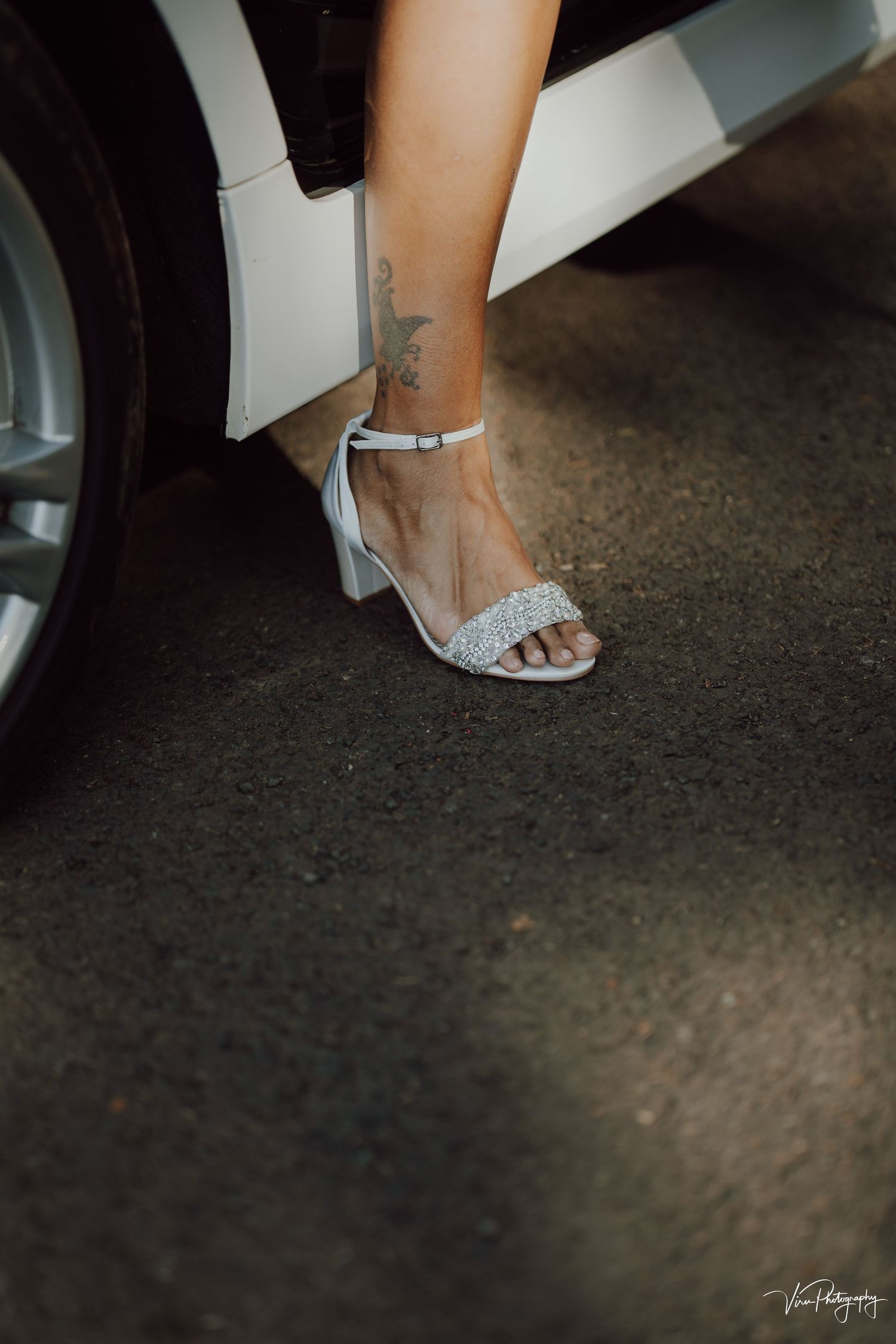 A foot wearing a sparkly silver ankle-strap sandal with a low block heel is shown next to a car door.