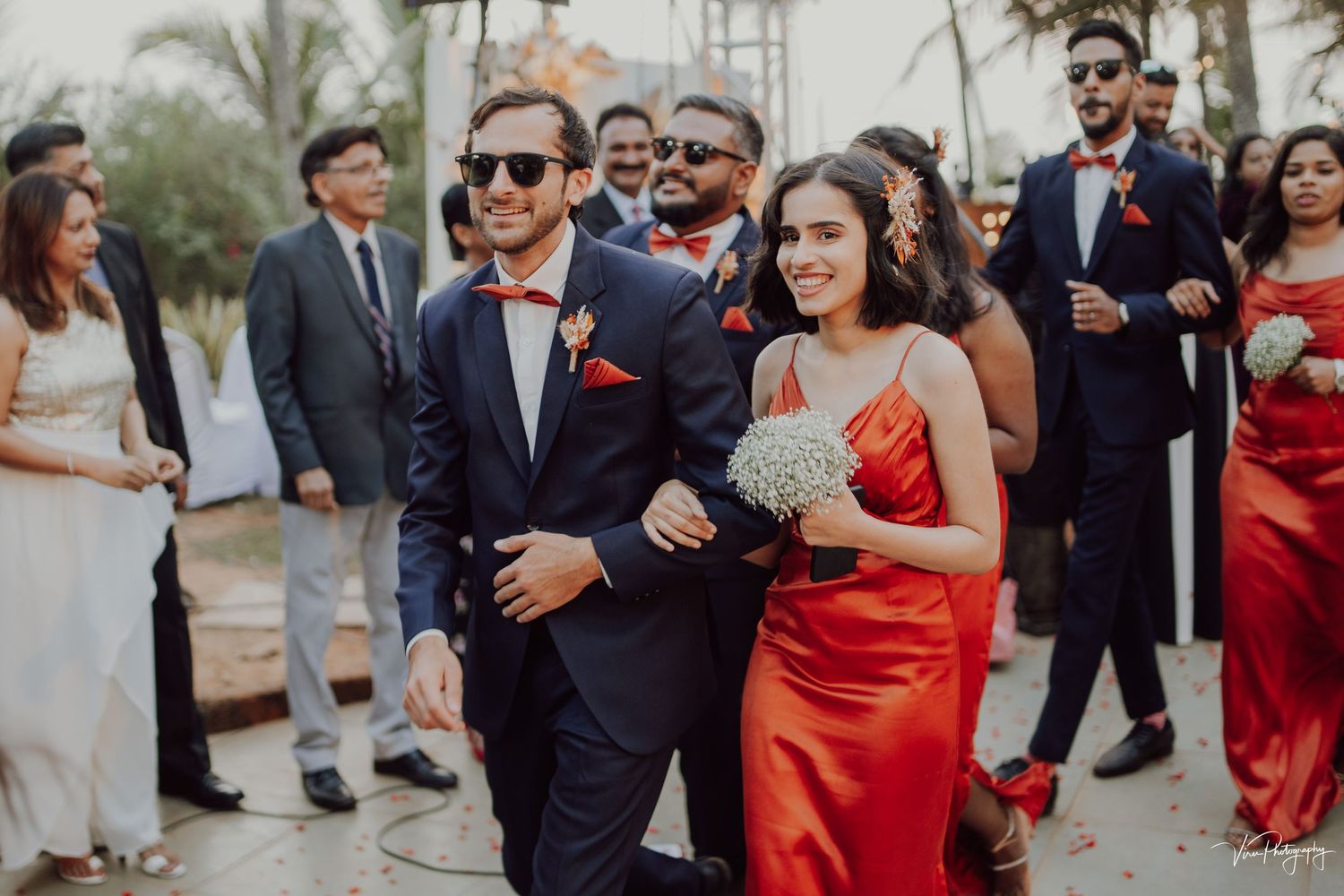 Bridal party members walk together outside with palm trees, featuring a vibrant red formal dress and black suits.