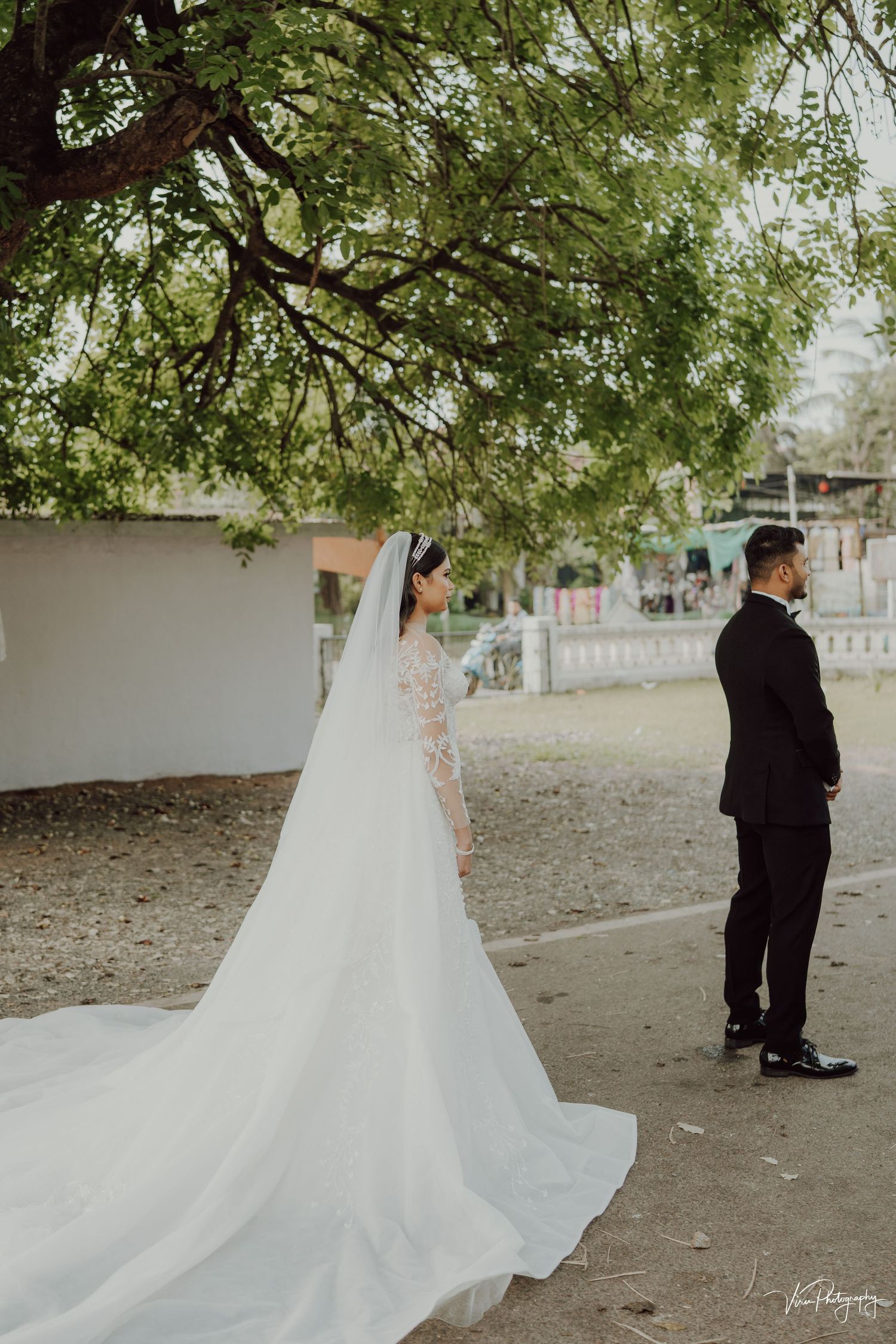 A couple stands outdoors during their wedding, with the bride in a long flowing veil gown and groom in classic tuxedo.