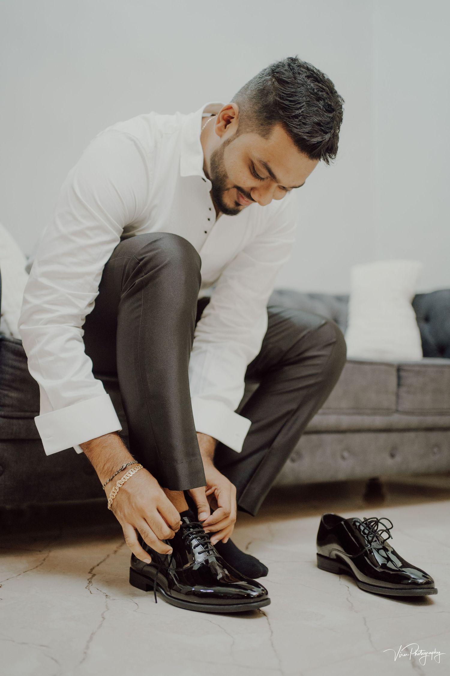 Person in formal attire sitting on gray couch tying shiny black dress shoes while getting ready.