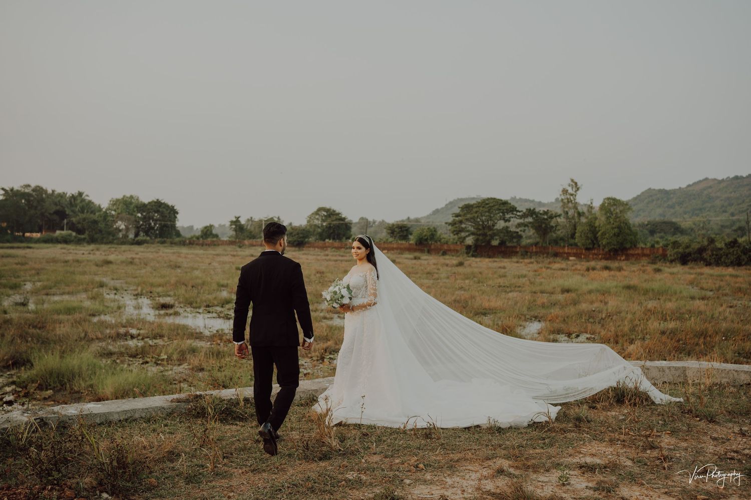 A couple strolls through a scenic field during their wedding photoshoot as the bride's long white veil flows in the breeze.