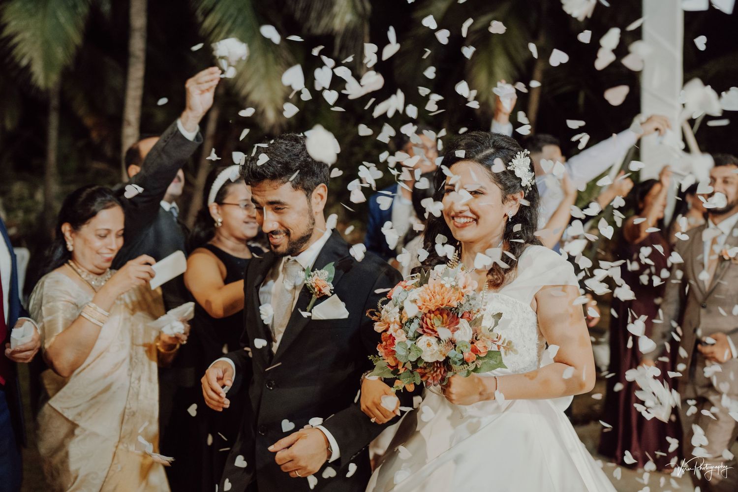 White flower petals float through the air as joyful newlyweds celebrate during an outdoor wedding ceremony.