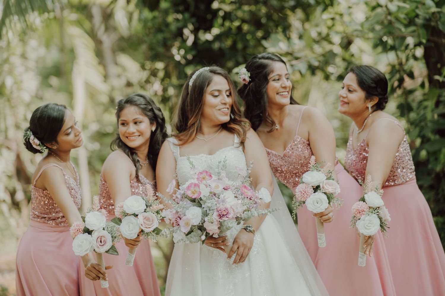 A bridal party in pink dresses and white gowns poses together while laughing in a garden.