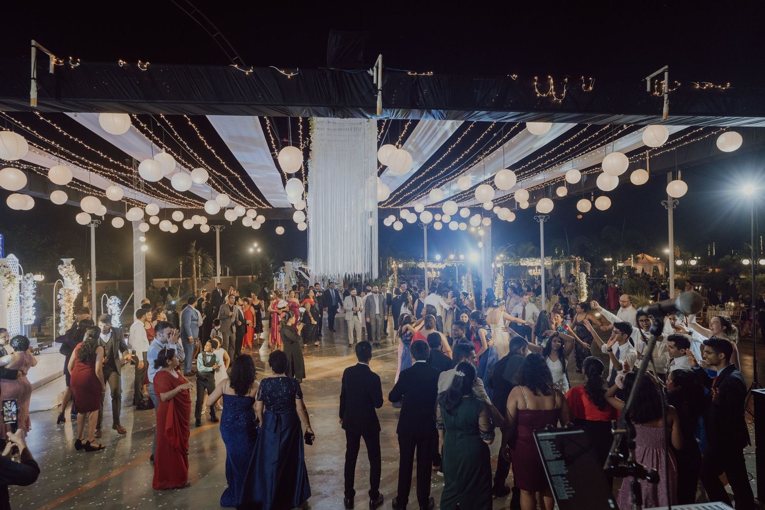 Large crowd dances under illuminated pavilion at wedding reception with festive lighting.