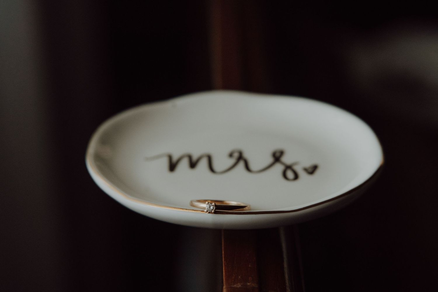 A wedding ring rests on a small white plate with 'mrs' written in cursive.