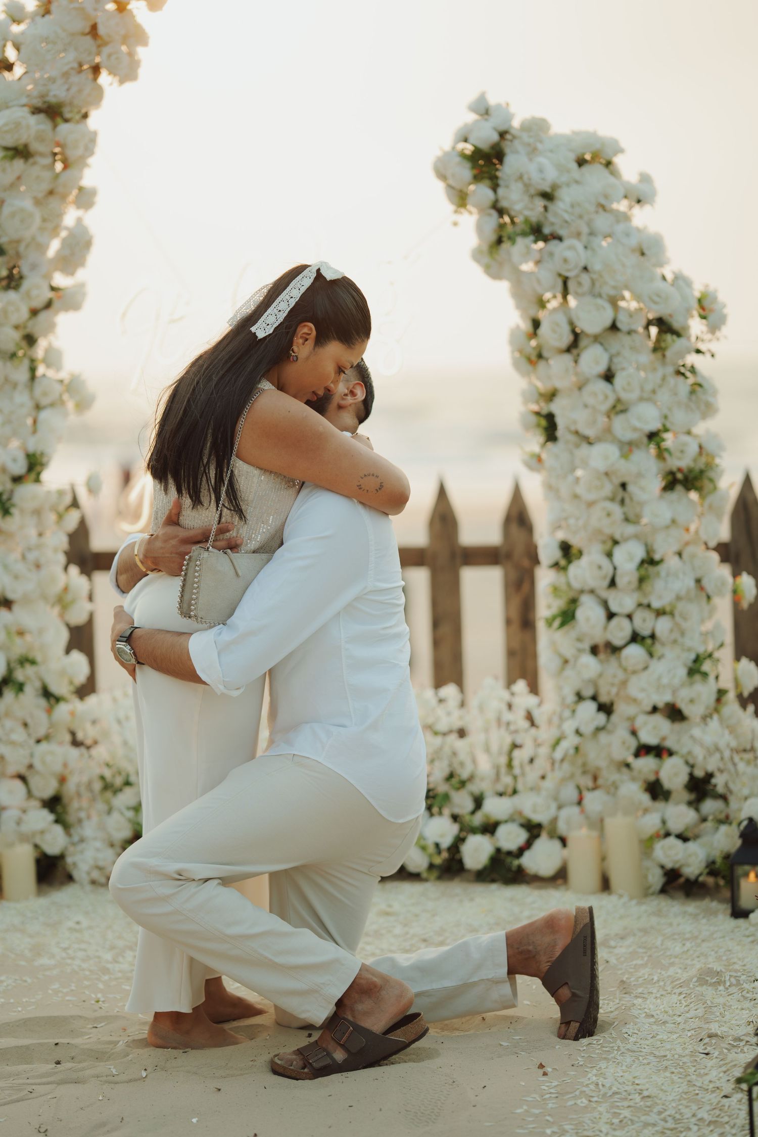 Couple embracing at a beach wedding ceremony with white floral arch and candles in the background.