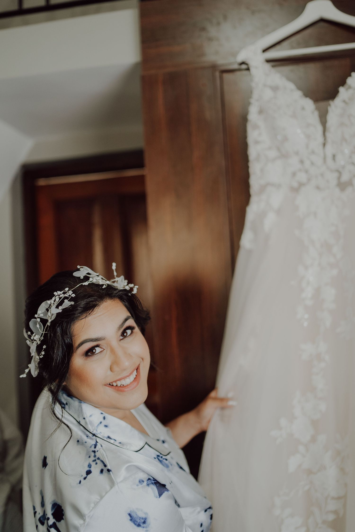 A delicate lace wedding dress hangs while someone in a floral robe admires it during pre-wedding preparations.