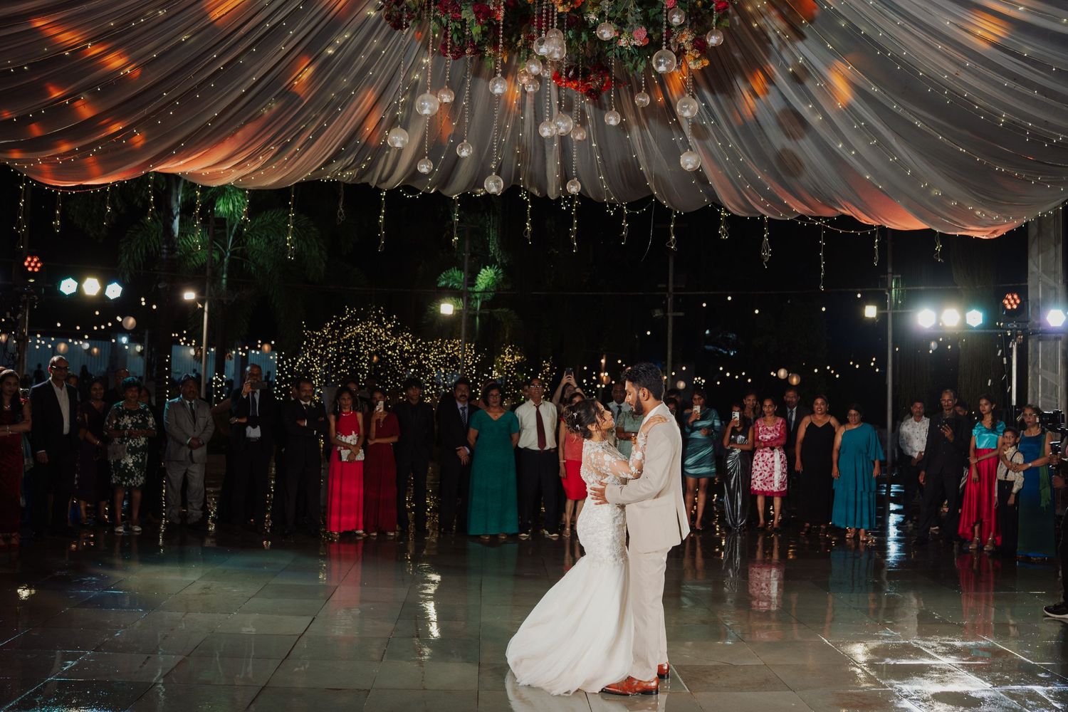 Bride and groom share their first dance on a reflective floor beneath a draped canopy and string lights as wedding guests watch.