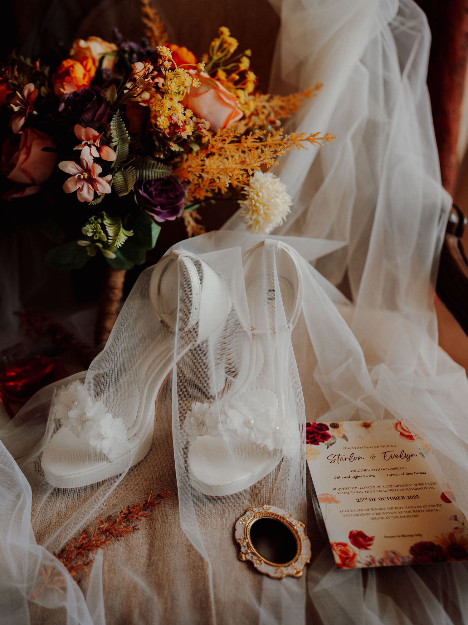 Bridal flat lay with white floral sandals, a sheer veil, an autumn-toned bouquet, a small gold mirror and a floral wedding invitation.