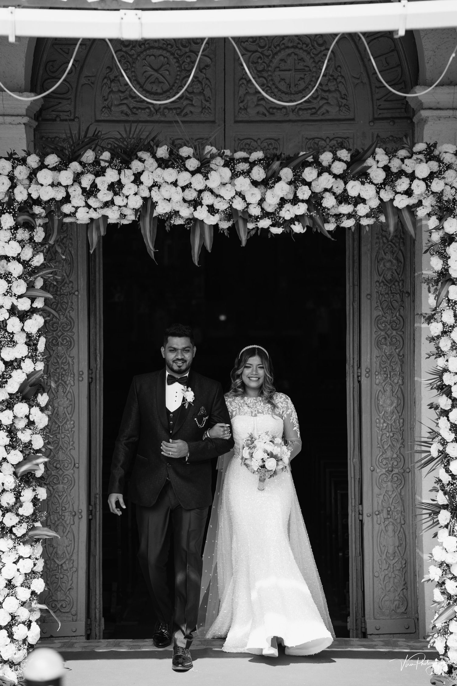 A wedding couple exits through an ornate doorway adorned with white floral arrangements in black and white photography.