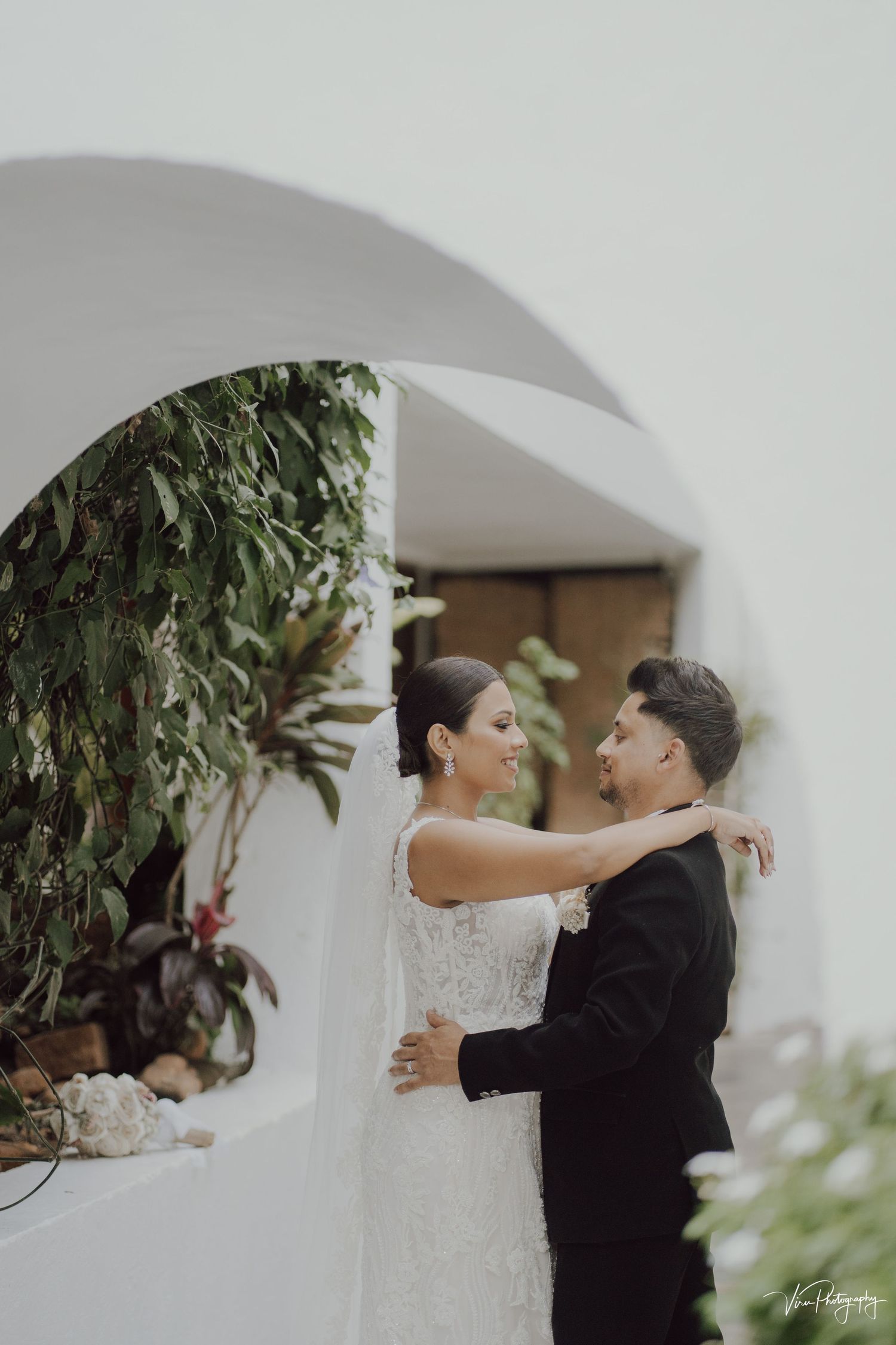 Newlywed couple embraces under white archway with green foliage at outdoor wedding venue.