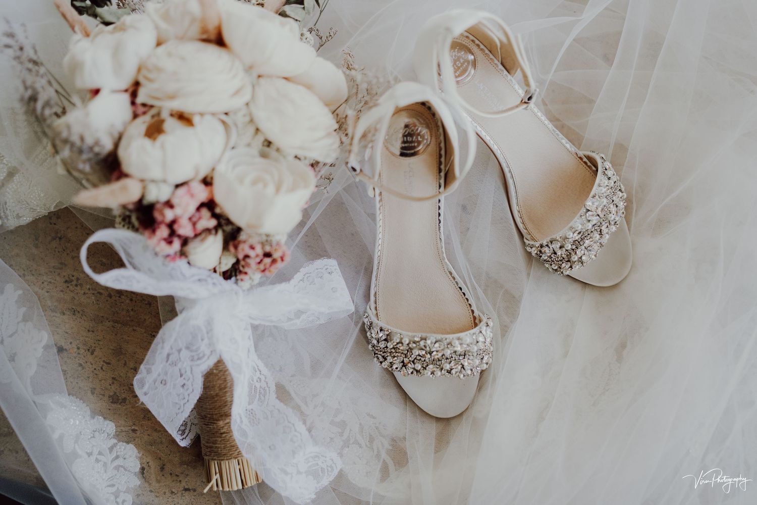 Elegant bridal shoes with sparkly embellishments displayed alongside white tulle veil and blush pink floral bouquet.