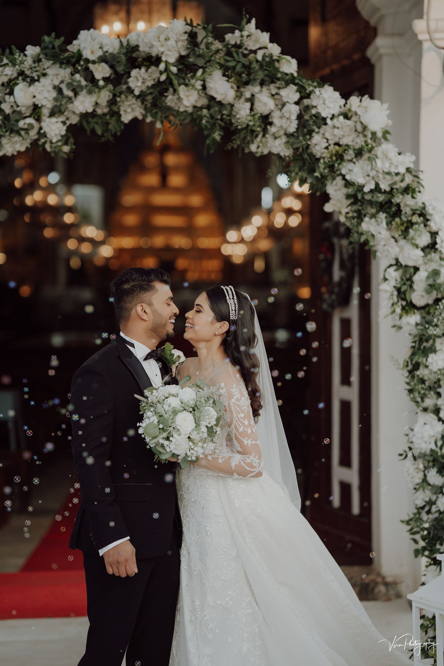 A couple shares an intimate moment under an elegant white floral arch with twinkling lights in a romantic wedding setting.