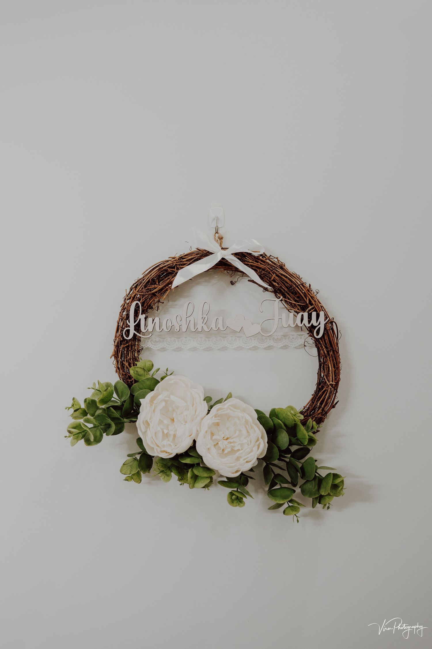 A minimalist grapevine wreath decorated with white roses and green foliage against a light gray wall.