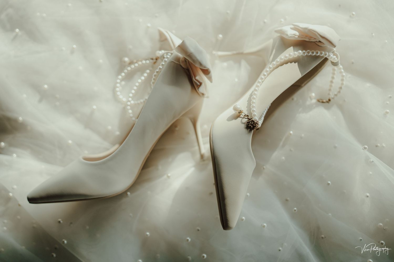 Elegant white satin bridal heels with pearl bow details rest on soft fabric in dreamy lighting.