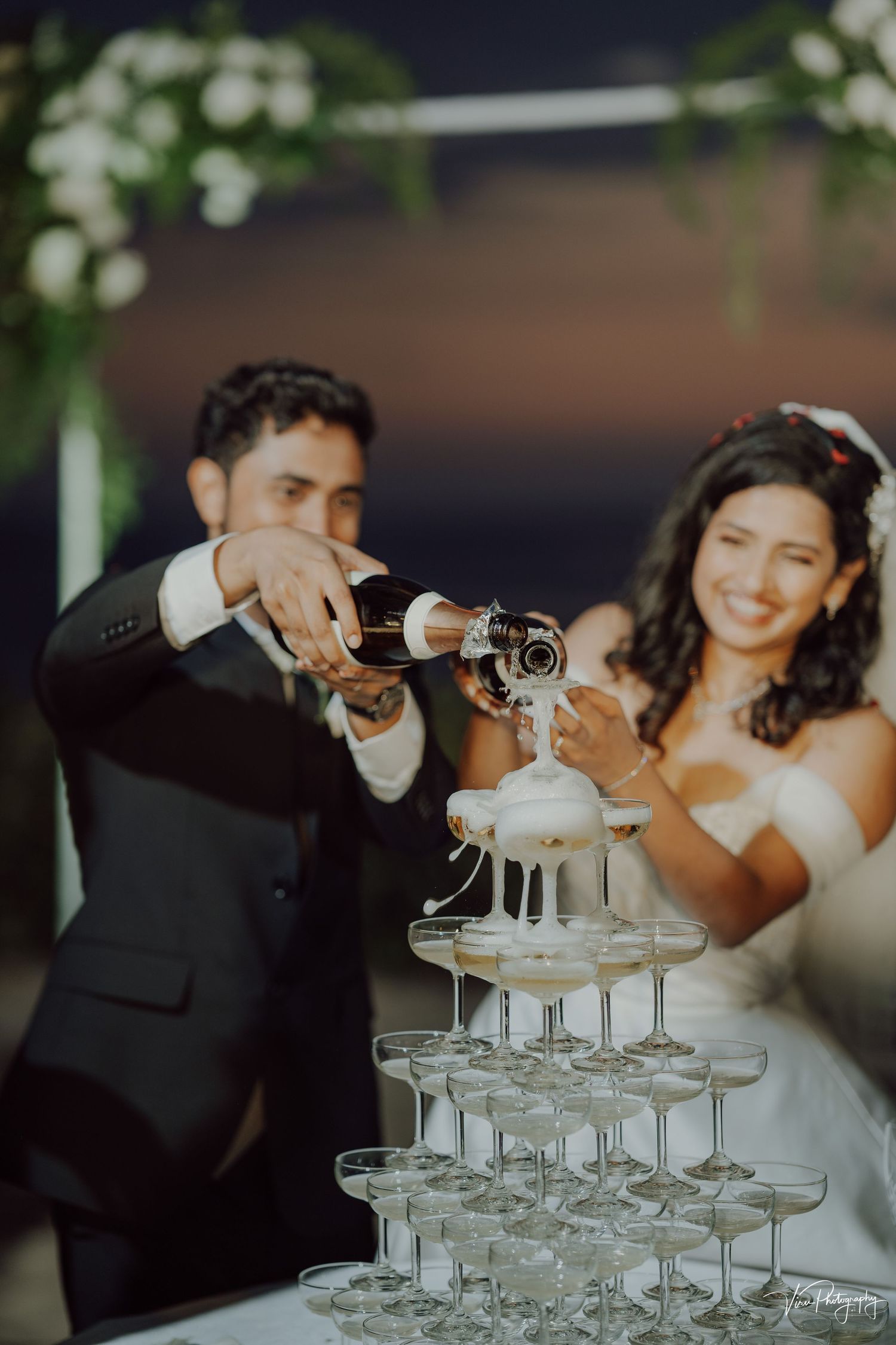 A couple pours champagne into a tower of stacked glasses during their wedding celebration.