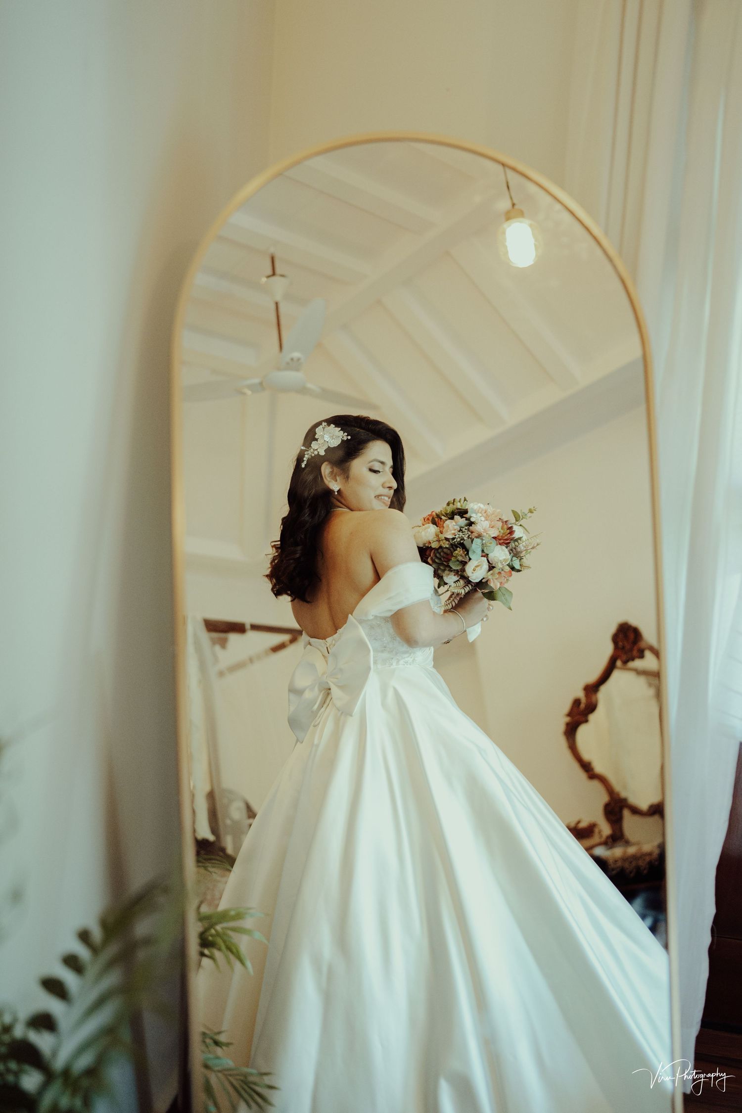 A bride in a flowing white wedding gown poses gracefully in front of an arched mirror holding a pink and white bouquet.