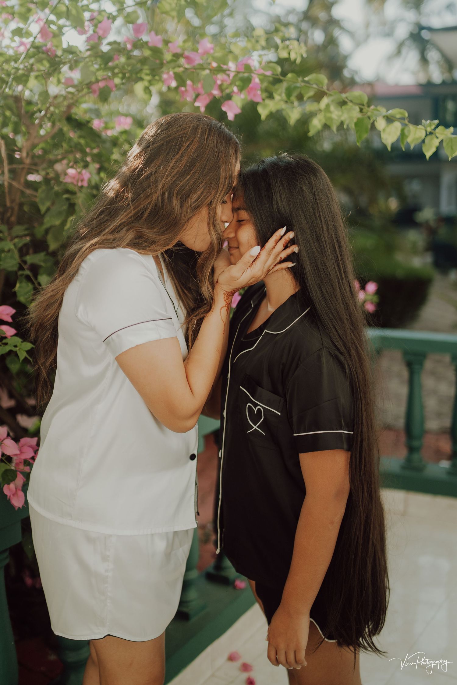 Two people sharing an intimate moment outdoors near pink flowers and greenery on a sunny day.