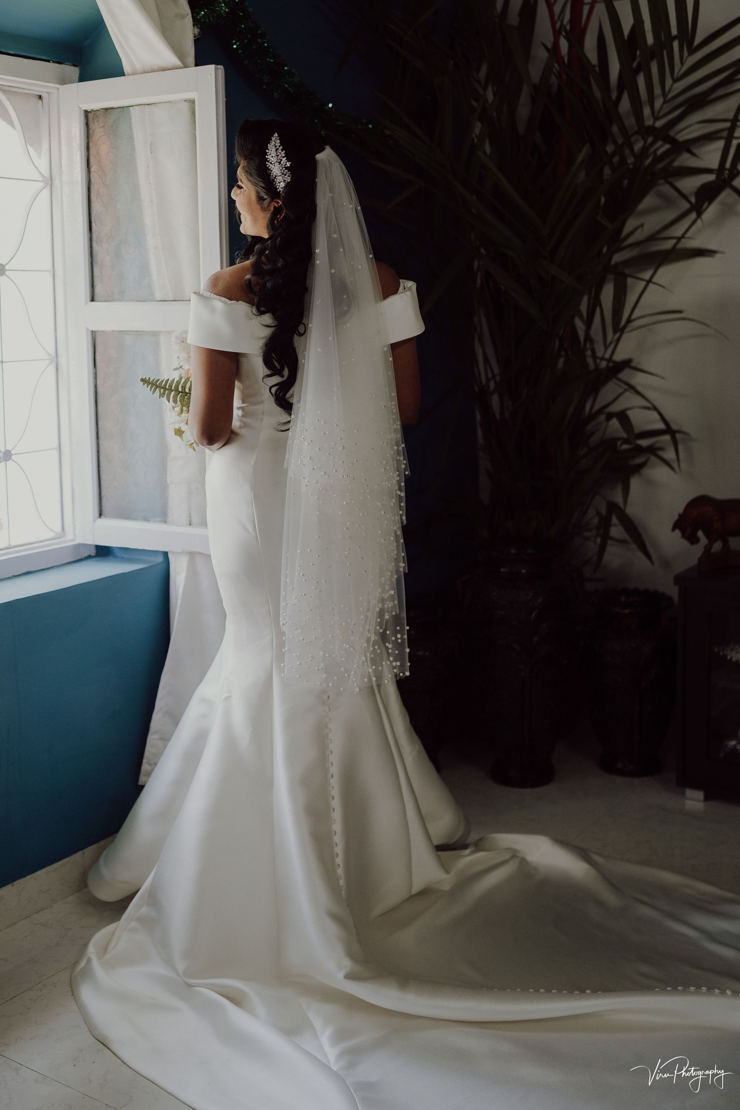 A bride in an elegant white mermaid wedding gown with a flowing veil stands by a window in a modern interior setting.