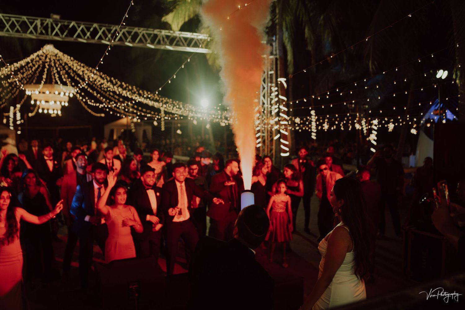 A crowd of people dance under string lights at a nighttime event with moody red lighting and atmospheric fog.