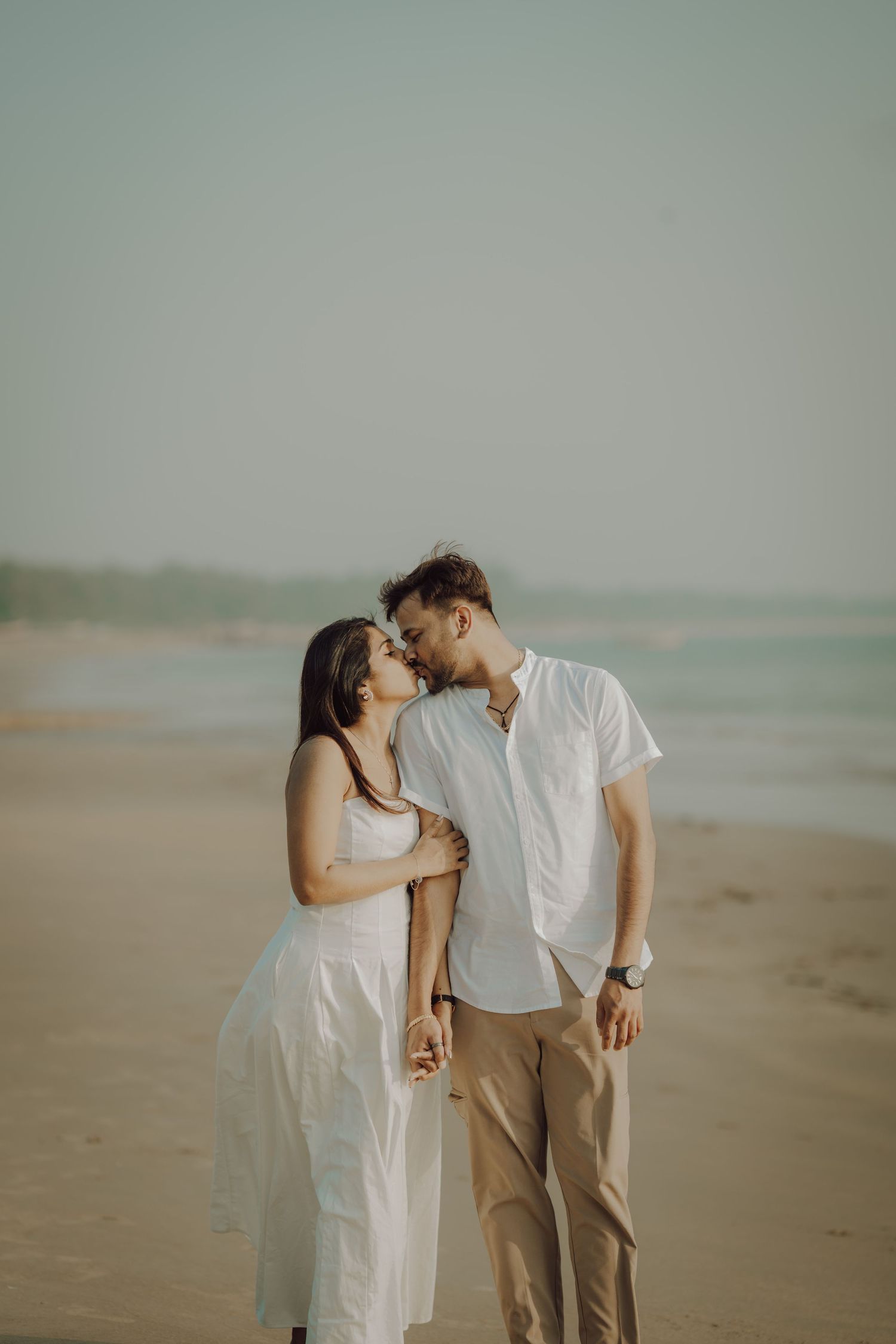 Romantic couple in light colored outfits share tender moments while walking along a misty beach at sunset.