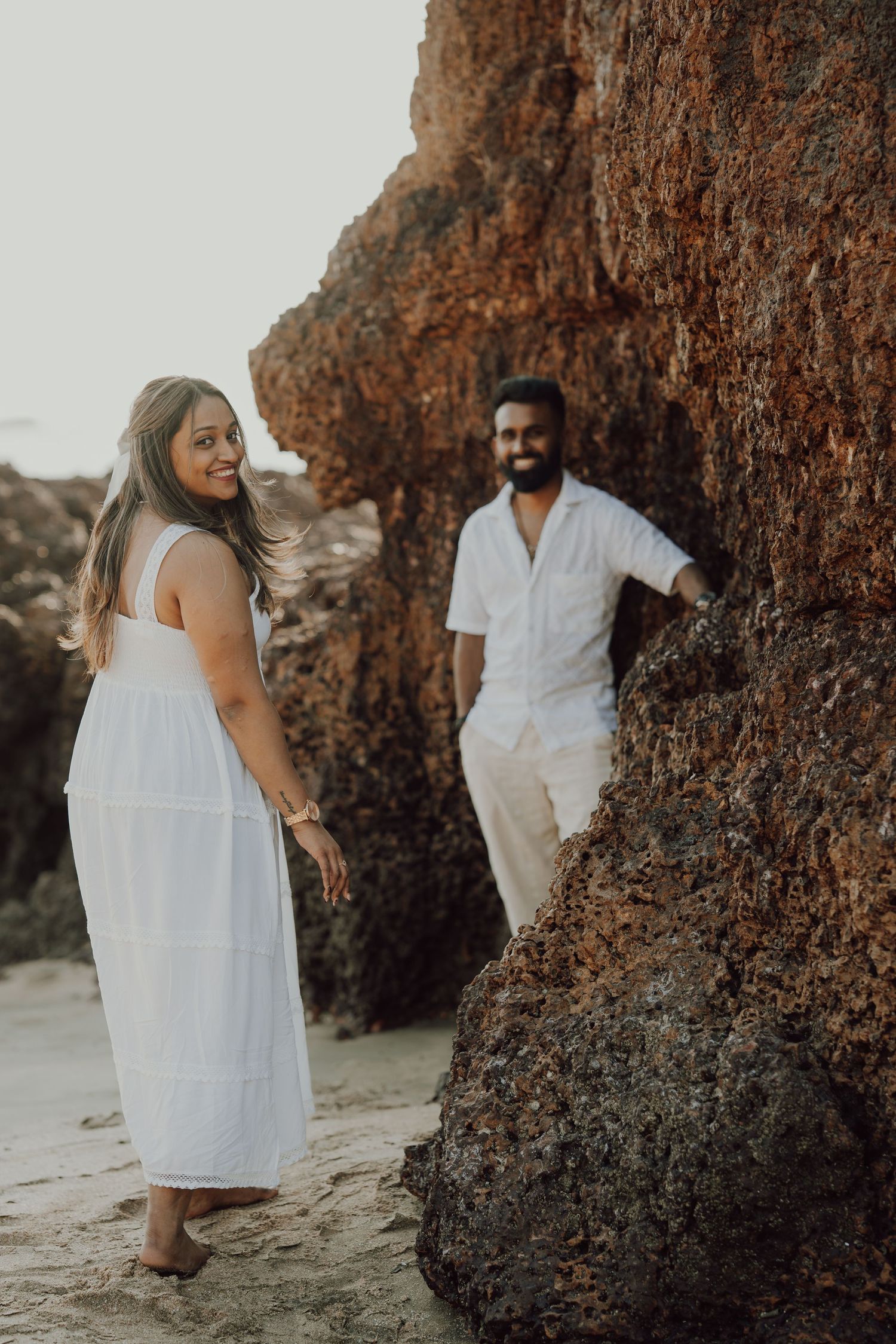 A couple in white outfits poses together near rocky coastal cliffs during sunset for romantic beach photos.