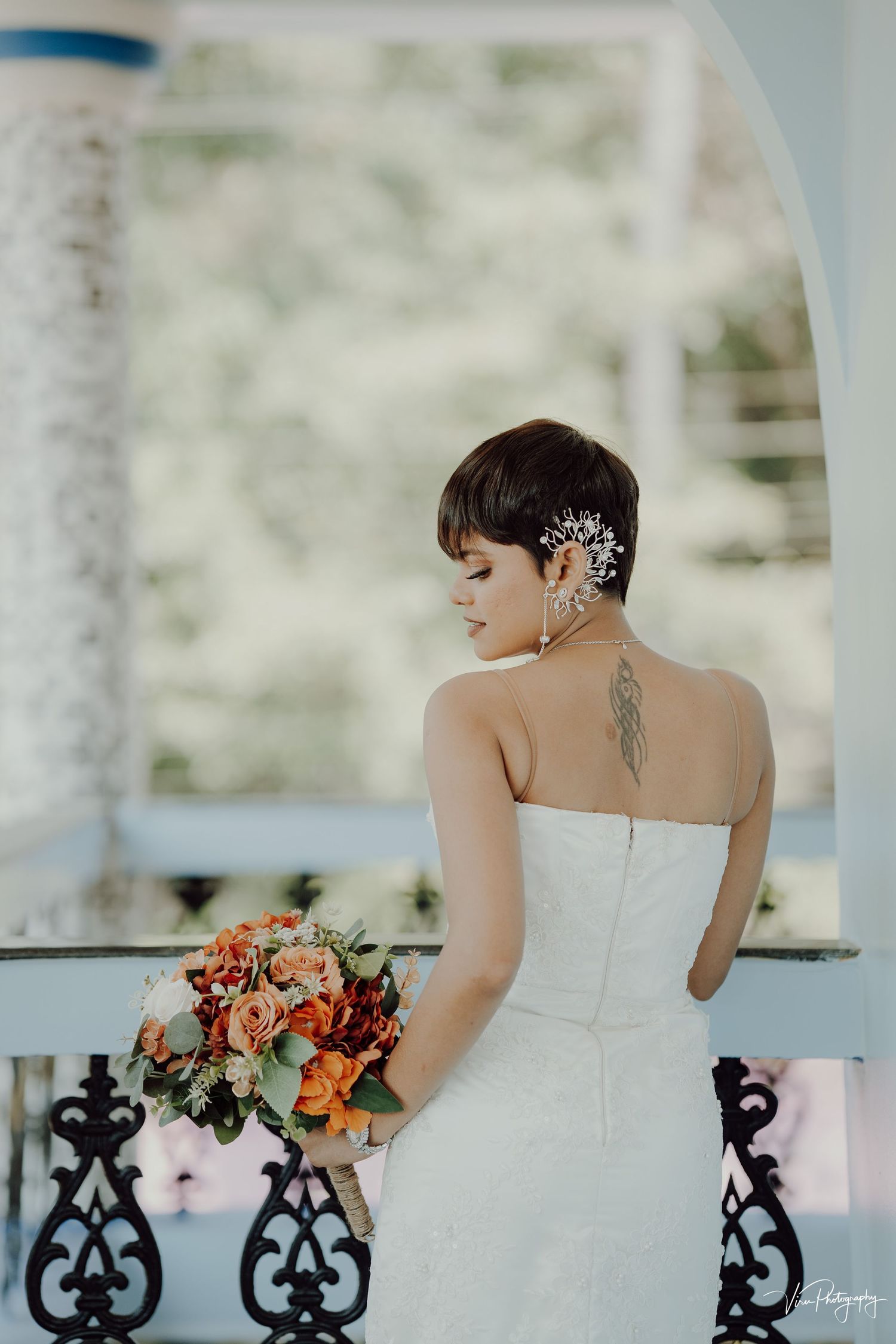 Back view of person in white wedding dress with pixie haircut and hair accessory holding coral bouquet.