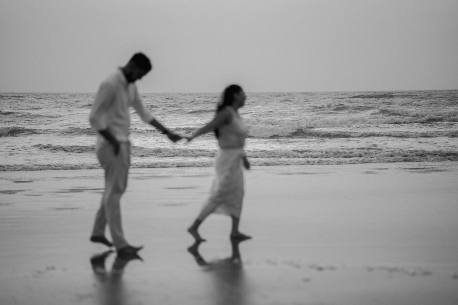 Black and white photo of a couple holding hands and walking barefoot on a beach by the ocean.
