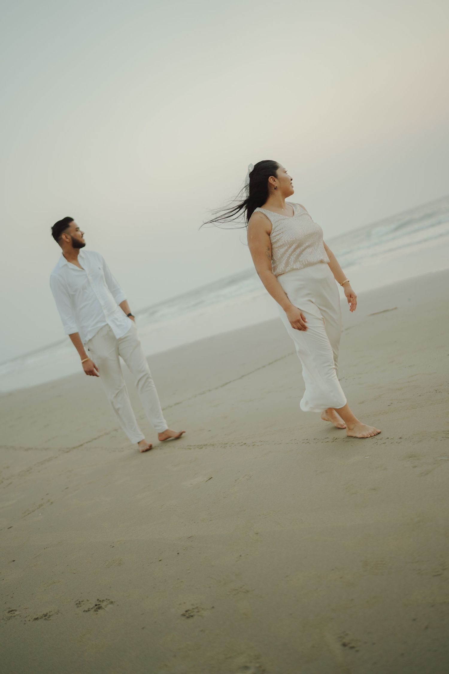 Couple in white outfits walking barefoot on a serene beach at dusk, woman with flowing hair looking up.