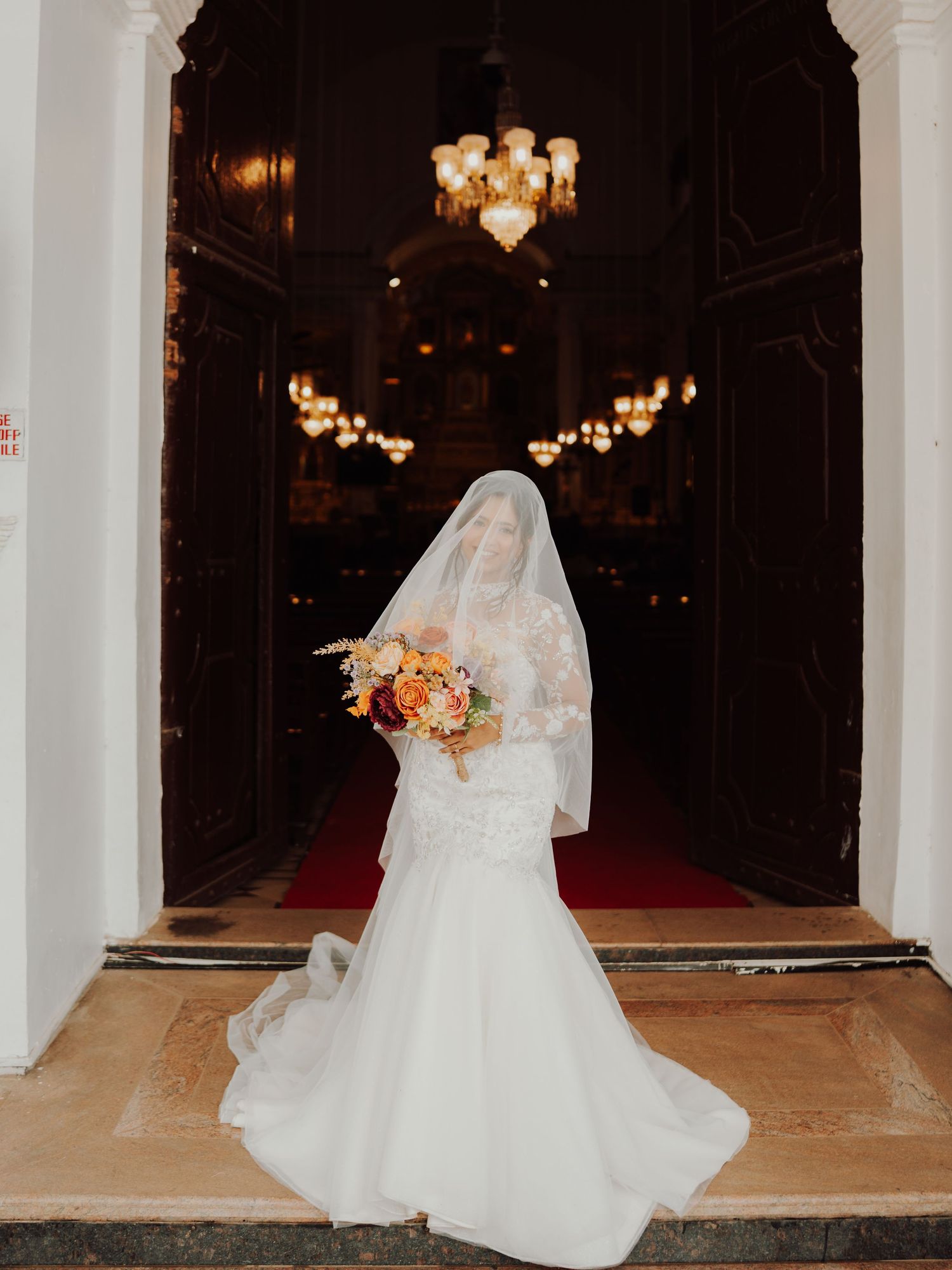 A bride in a white mermaid gown and sheer veil holds an orange bouquet at the doorway of a grand church with chandeliers.