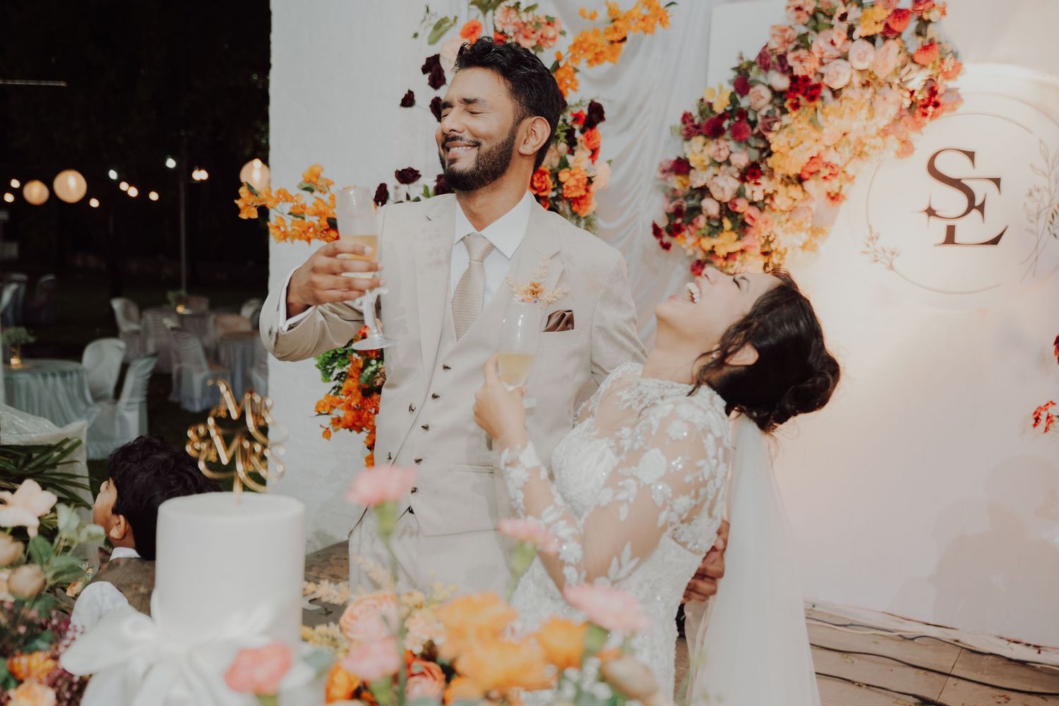A groom in a beige suit celebrates with his bride beside a wedding cake and colorful floral decor at an evening reception.