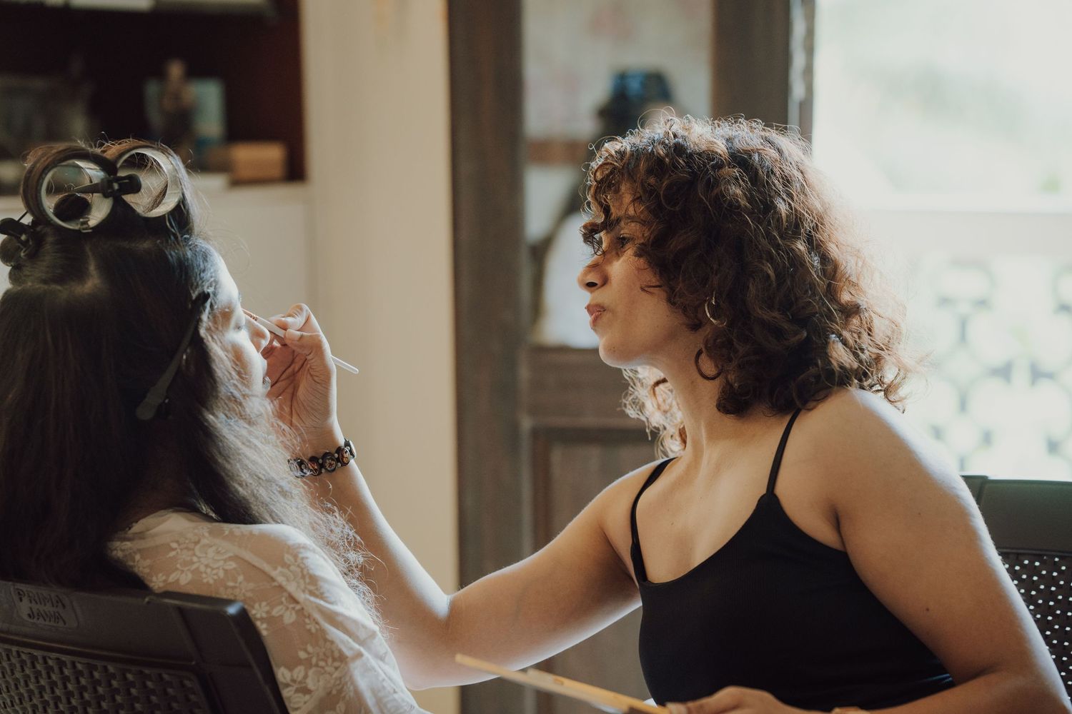 A makeup artist with curly dark hair applies makeup to a seated woman with hair in vintage pin curls in a sunlit room.