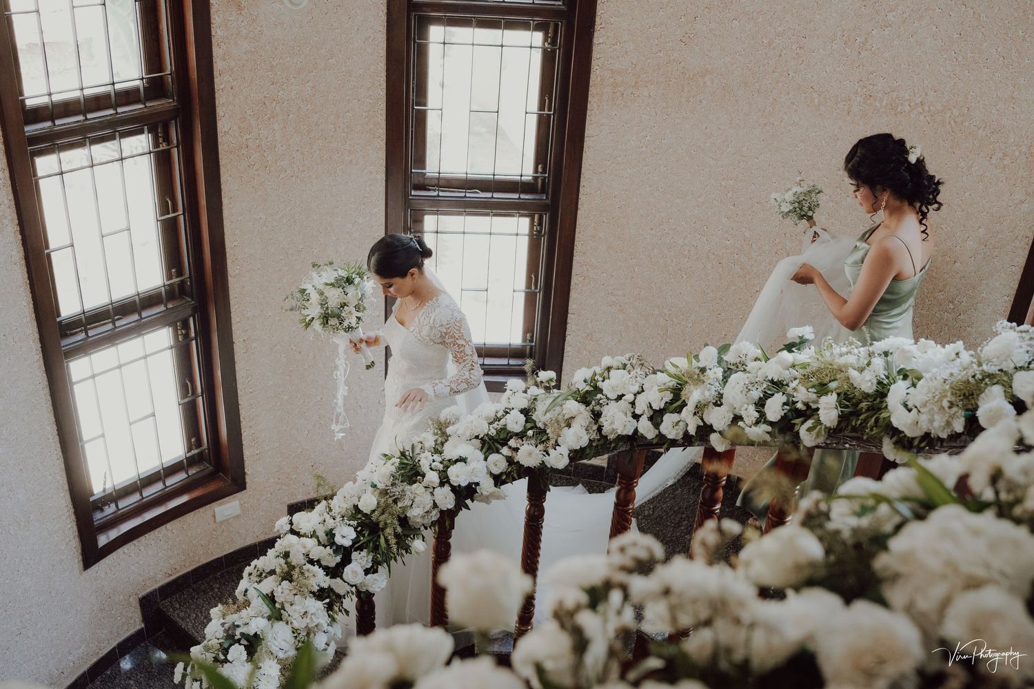 Two figures surrounded by elegant white floral arrangements in a classic room with tall windows.