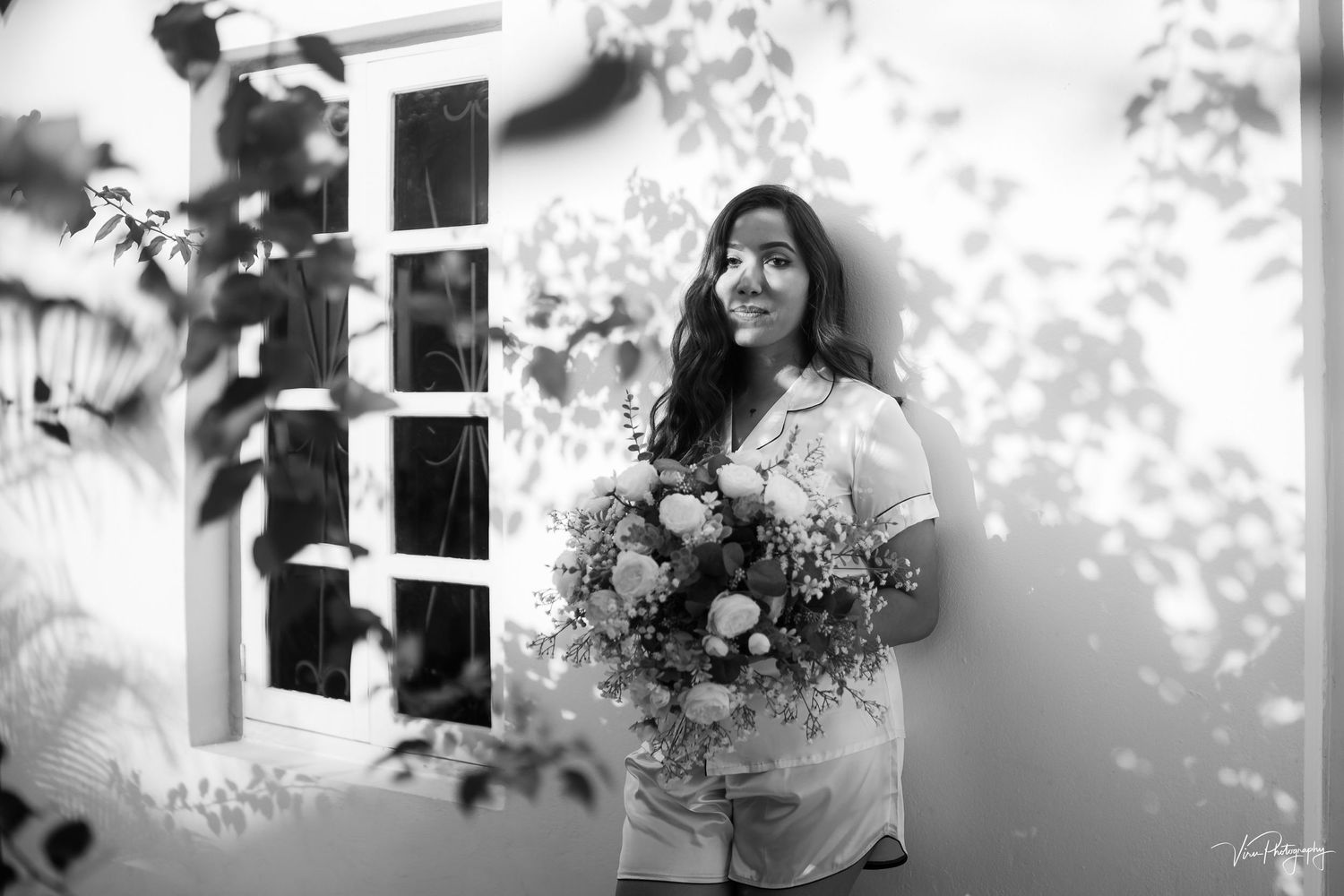 A person in casual attire holds a lush bouquet of flowers while standing near a window in a black and white photograph.