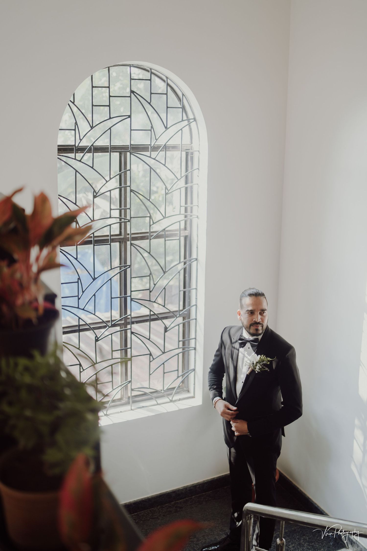 Art deco stained glass window frames a stylish figure in formal attire standing near a potted plant in bright interior space.